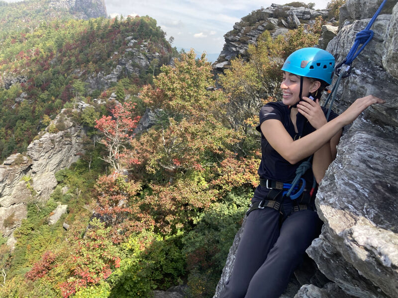 A woman wearing a blue helmet is rock climbing on a sunny day. She is attached to a rope and is looking to her right. The background features a mountain range with trees in autumn colors. The rock face she is climbing is light gray and textured.